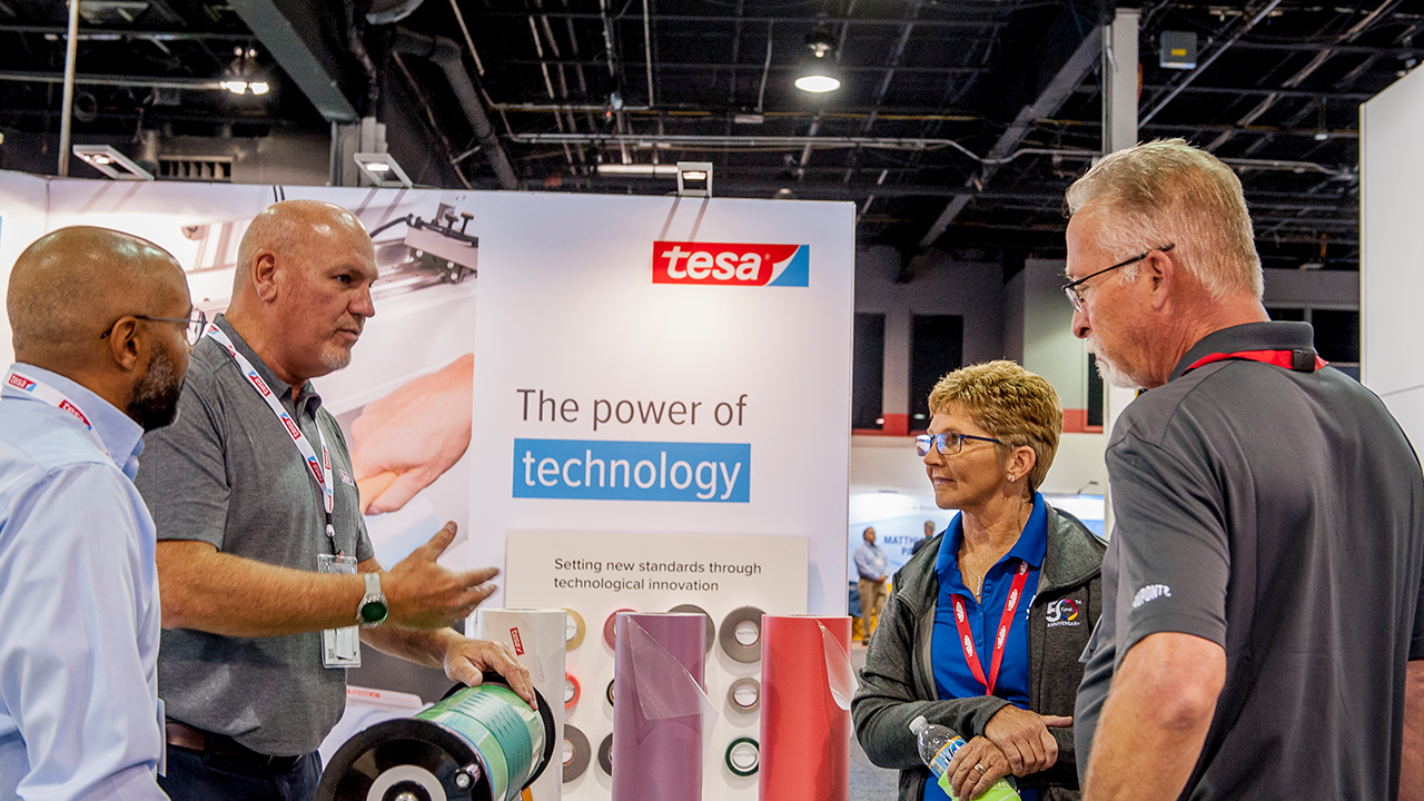Four people look at a self-adhesive sleeve on a cylinder at the Tesa Tapes booth